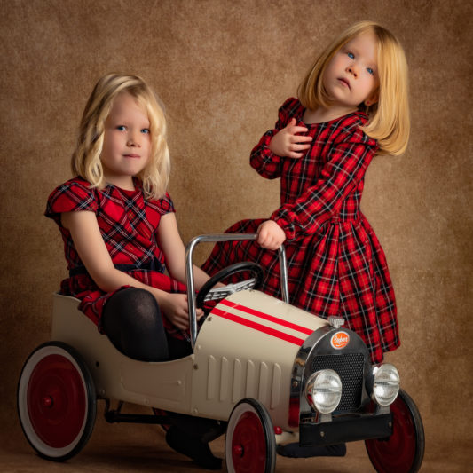 portrait fine-art de 2 sœurs habillées avec robe à carreaux rouge et noir et la voiture à pédale