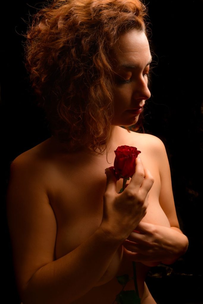 femme nue lumière douce cheveux bouclés portant une rose rouge sur son cœur.