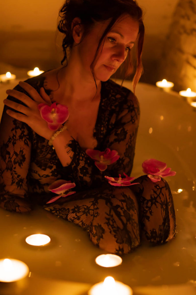 femme avec robe en dentelle dans un bain éclairée avec de nombreuses bougies posées au bord de la baignoire et des fleurs sur elle