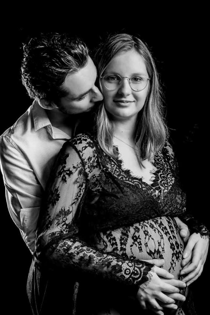 photo studio grossesse femme enceinte vêtue d'une robe en dentelle noire dans les bras de son mari en noir et blanc