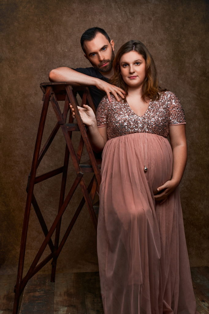 photo studio grossesse femme enceinte vêtue d'une robe en tulle rose avec son mari accoudé à un vieil escabeau en bois