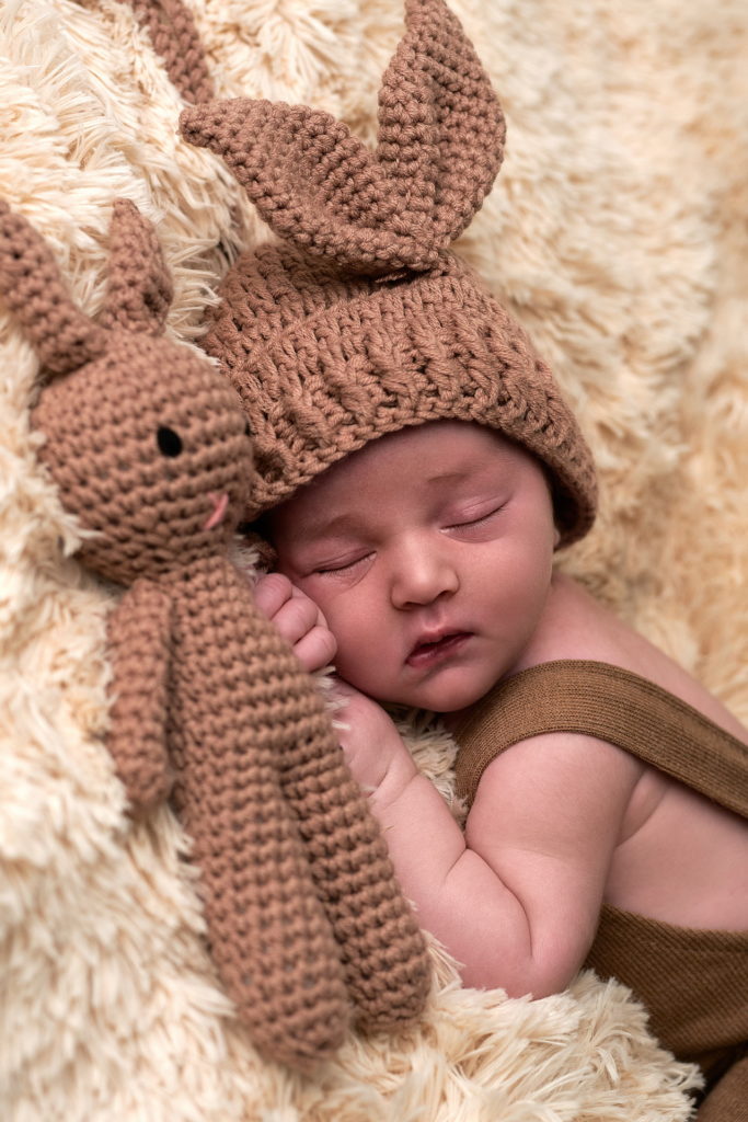 bébé endormi sur une couverture très douce, il est porte une salopette marron, porte un bonnet marron et fait un câlin à son doudou