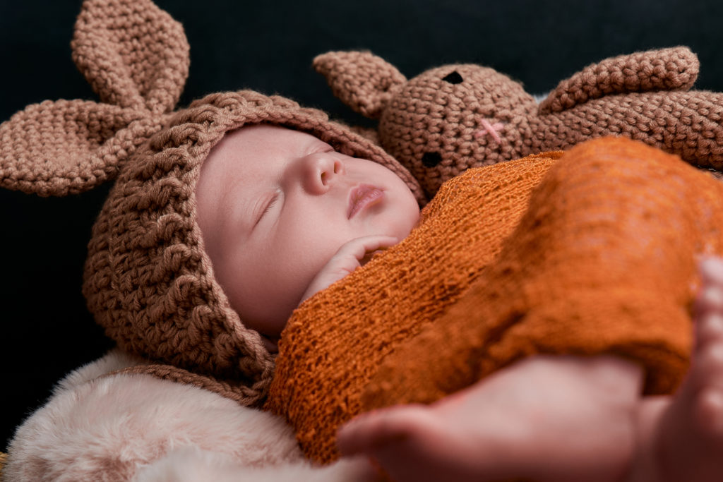 bébé endormi sur une couverture très douce, il est emmailloté dans un linge orange, porte un bonnet marron et fait un câlin à son doudou
