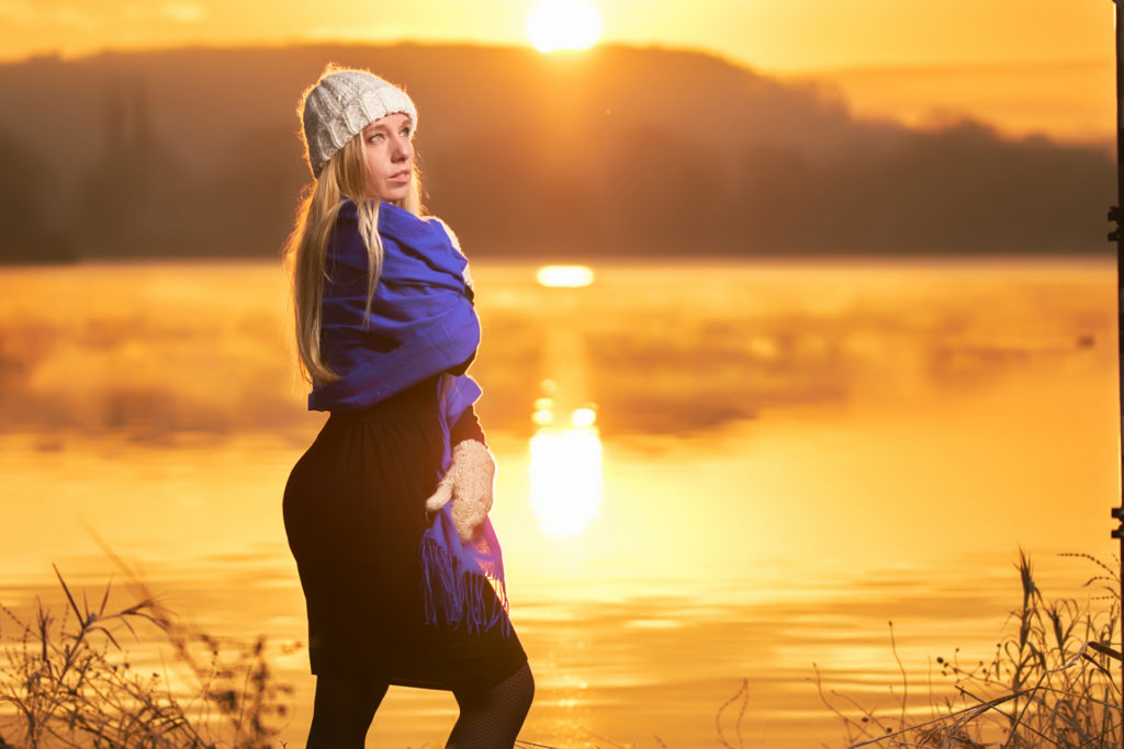 portrait d'une femme au bord d'un lac l’hiver pendant un levé de soleil portant un bonnet gris et une écharpe bleue