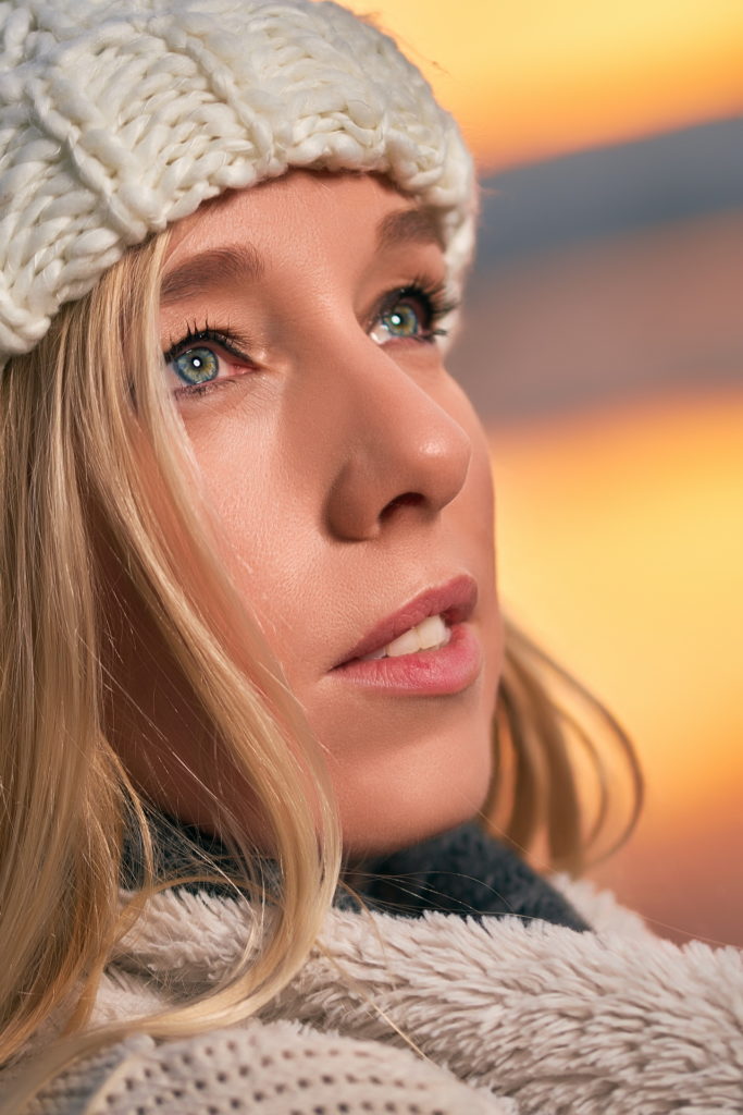 portrait gros plan d'une femme au bord d'un lac l’hiver pendant un levé de soleil portant un bonnet et un manteau épais